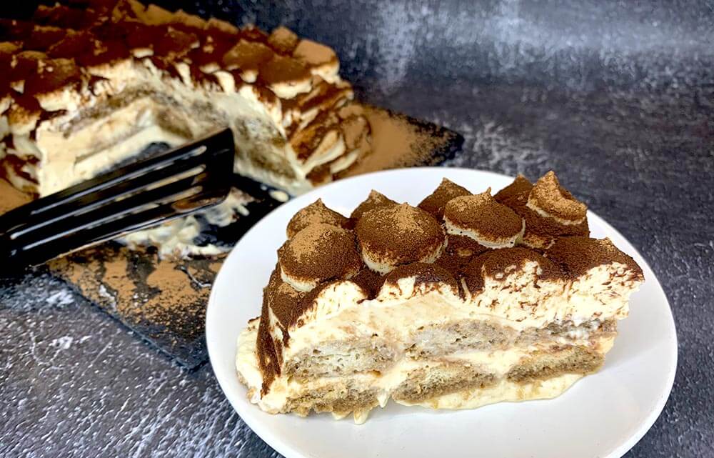 Detail rozkrojeného tiramisu na bielom tanieriku, v pozadí tiramisu na bridlicovej tácke